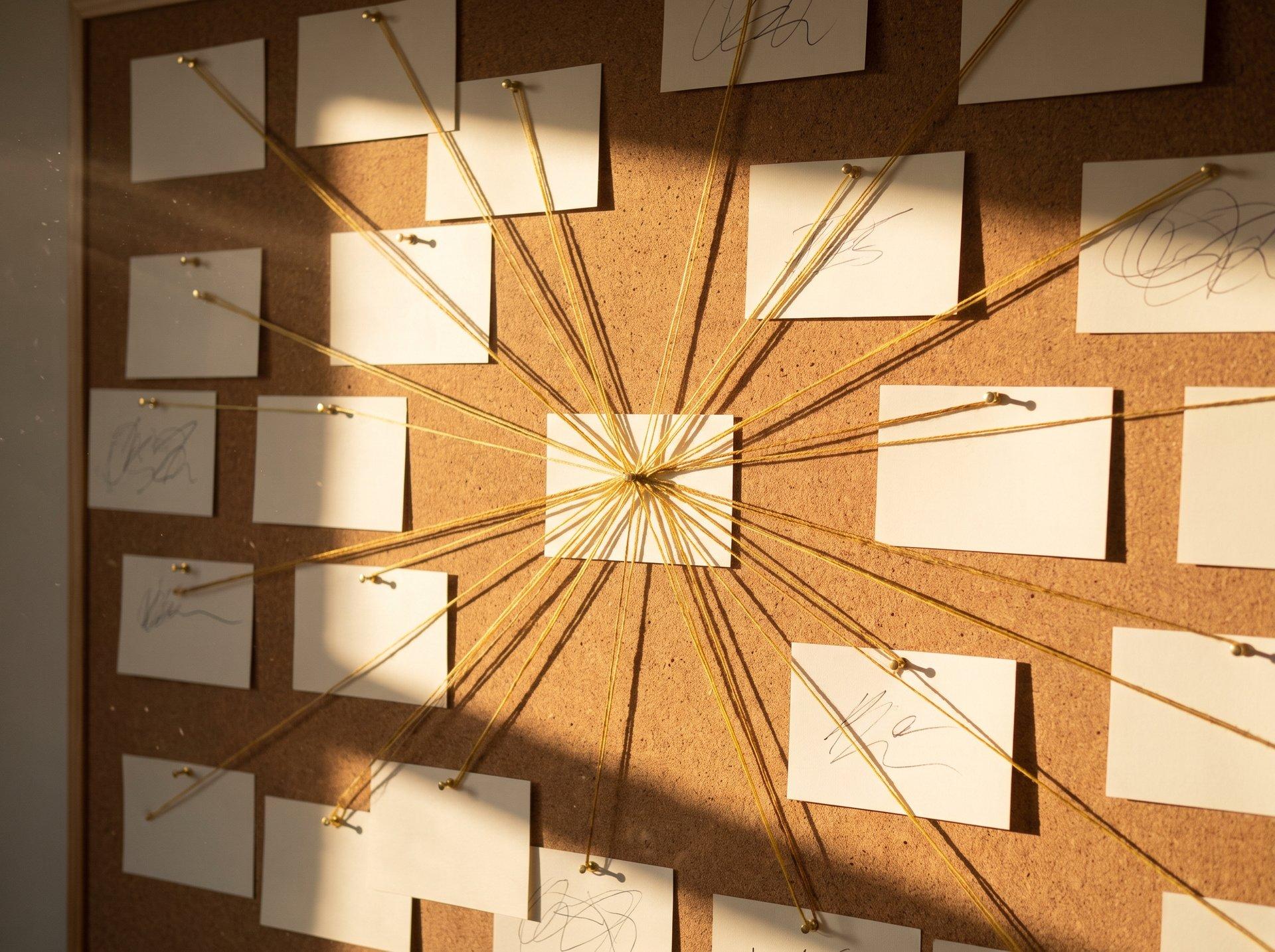 Index cards pinned to a cork wall, joined by thin gold thread, lit from one side