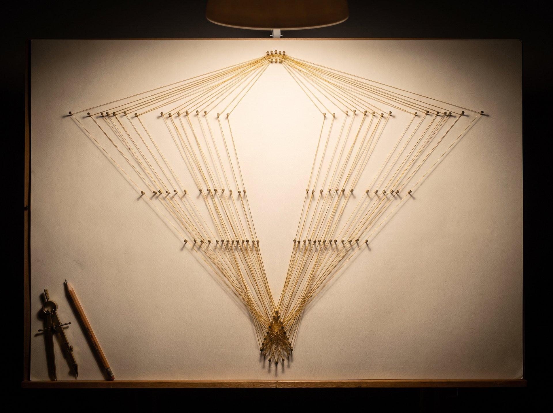 An overhead view of a drafting table with brass pins and thread forming a branching pattern